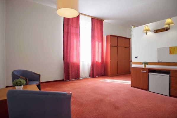A room with a wooden desk, a chair, a small refrigerator, a cabinet, and a large window with red curtains. - Hotel Selce (Crikvenica)