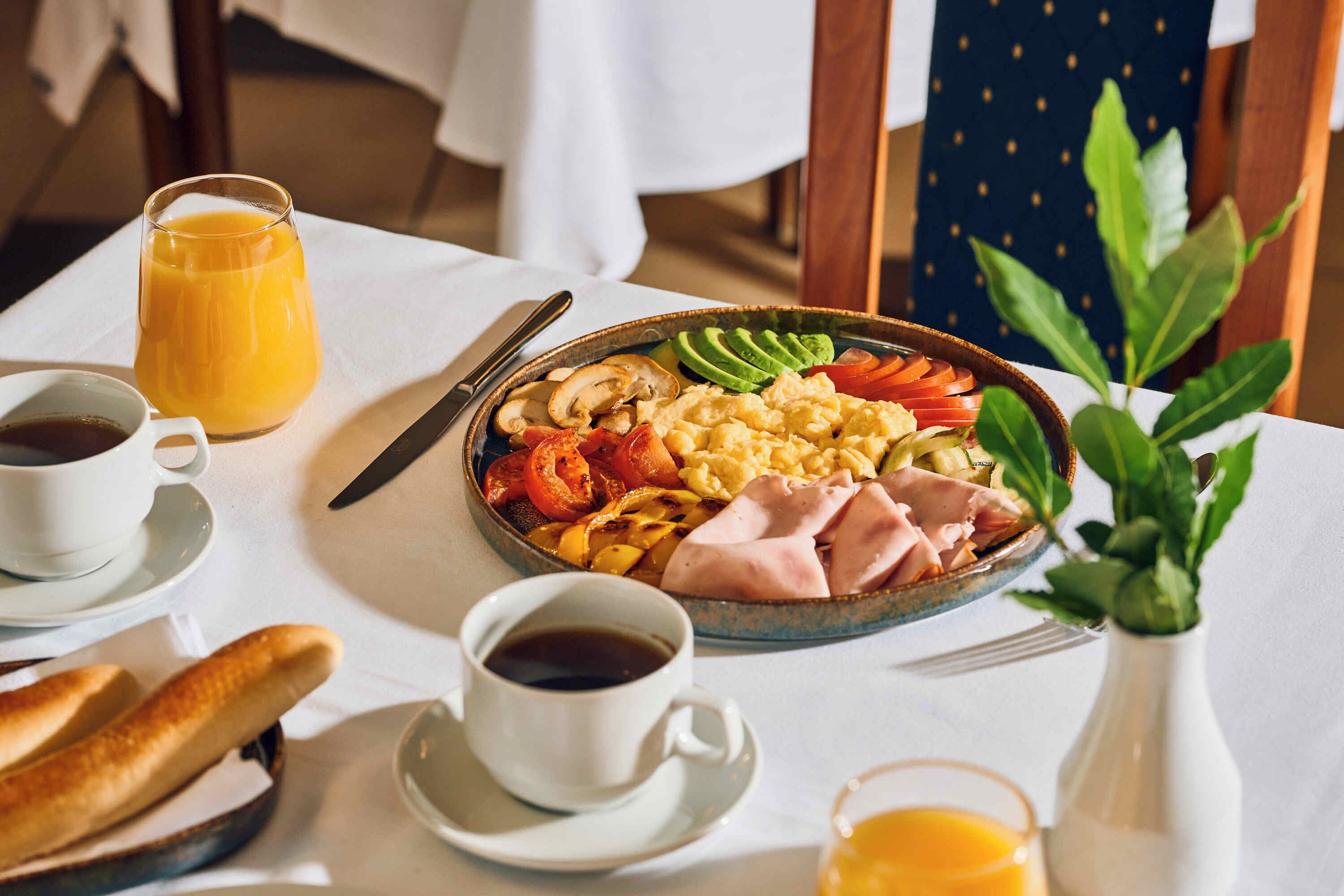 En frukostuppläggning med en tallrik innehållande äggröra, avokado, tomater, svamp och skinka, tillsammans med två koppar kaffe och ett glas apelsinjuice.