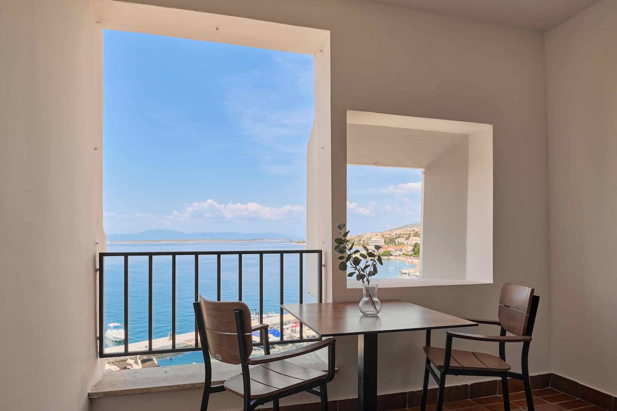 A balcony with a table and chairs, a vase with a plant, and a view of the sea and distant buildings.