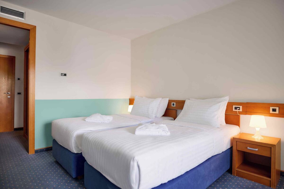 A hotel room with two beds, a wooden headboard, a bedside table with a lamp, and a carpeted floor.