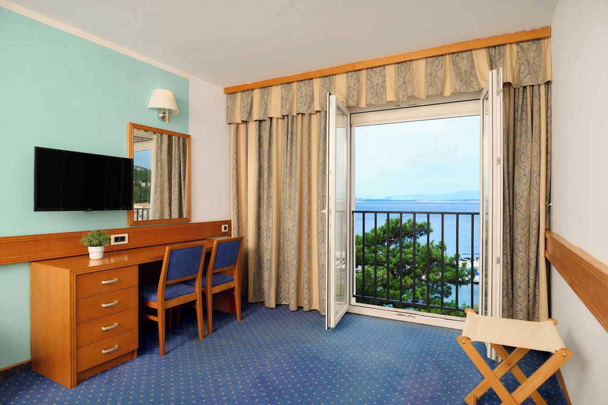 A hotel room with a wooden desk, a chair, a television, a balcony with a view of the sea, and a window with curtains.