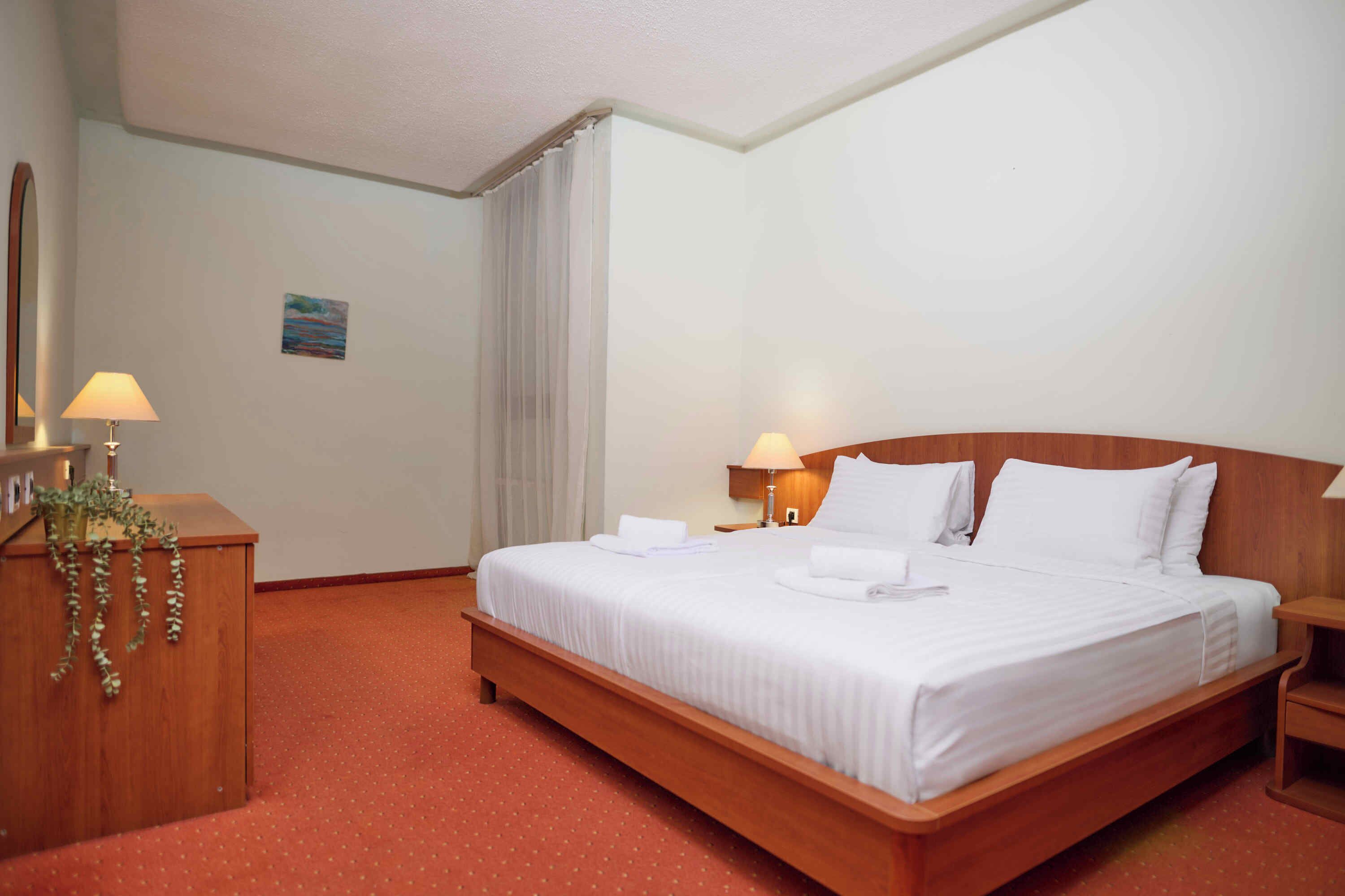 A hotel room with a large bed, two bedside tables, a desk with a lamp, and a small painting on the wall.