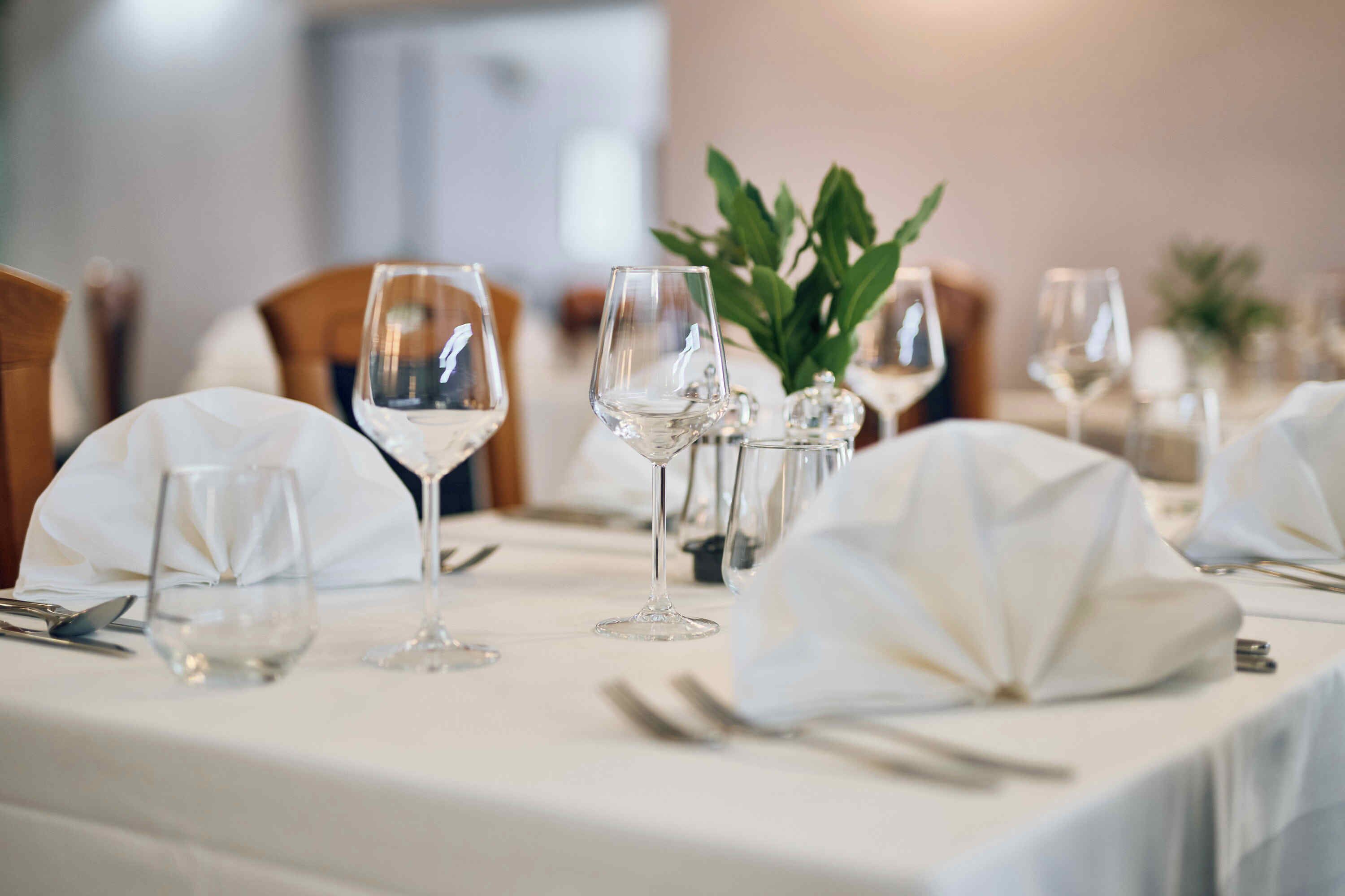A dining table set with white tablecloths, clear glassware, and a centerpiece of green foliage.