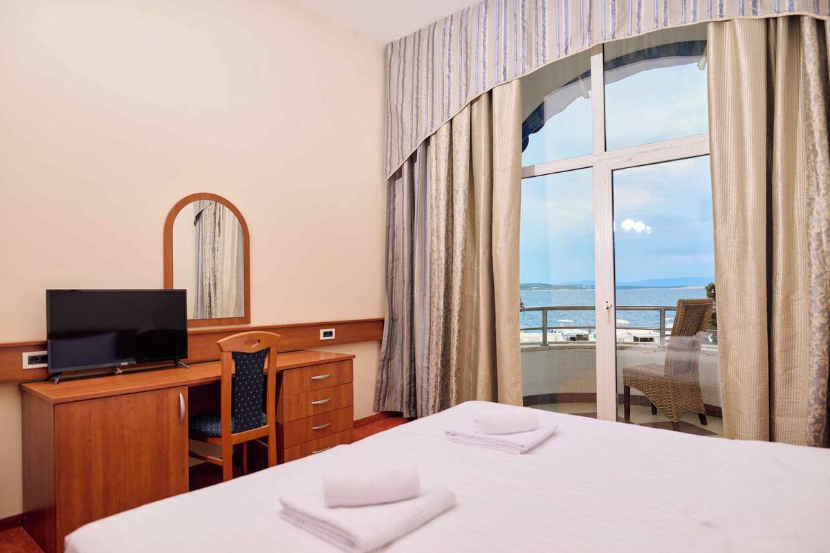 A hotel room with a bed, a desk with a television, a chair, and a view of the sea through a balcony.