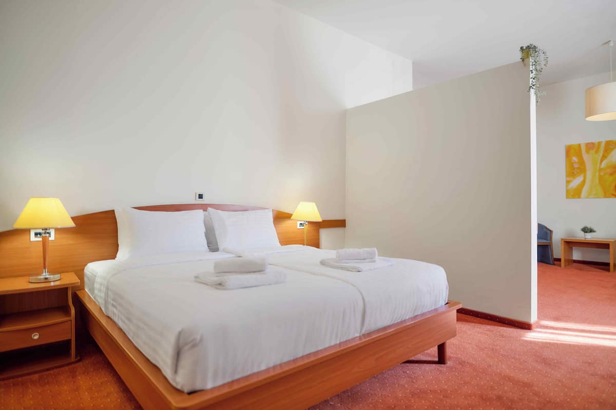 A neatly made double bed with white linens and two pillows, flanked by bedside tables with lamps, in a room with a wooden headboard and a red carpet.