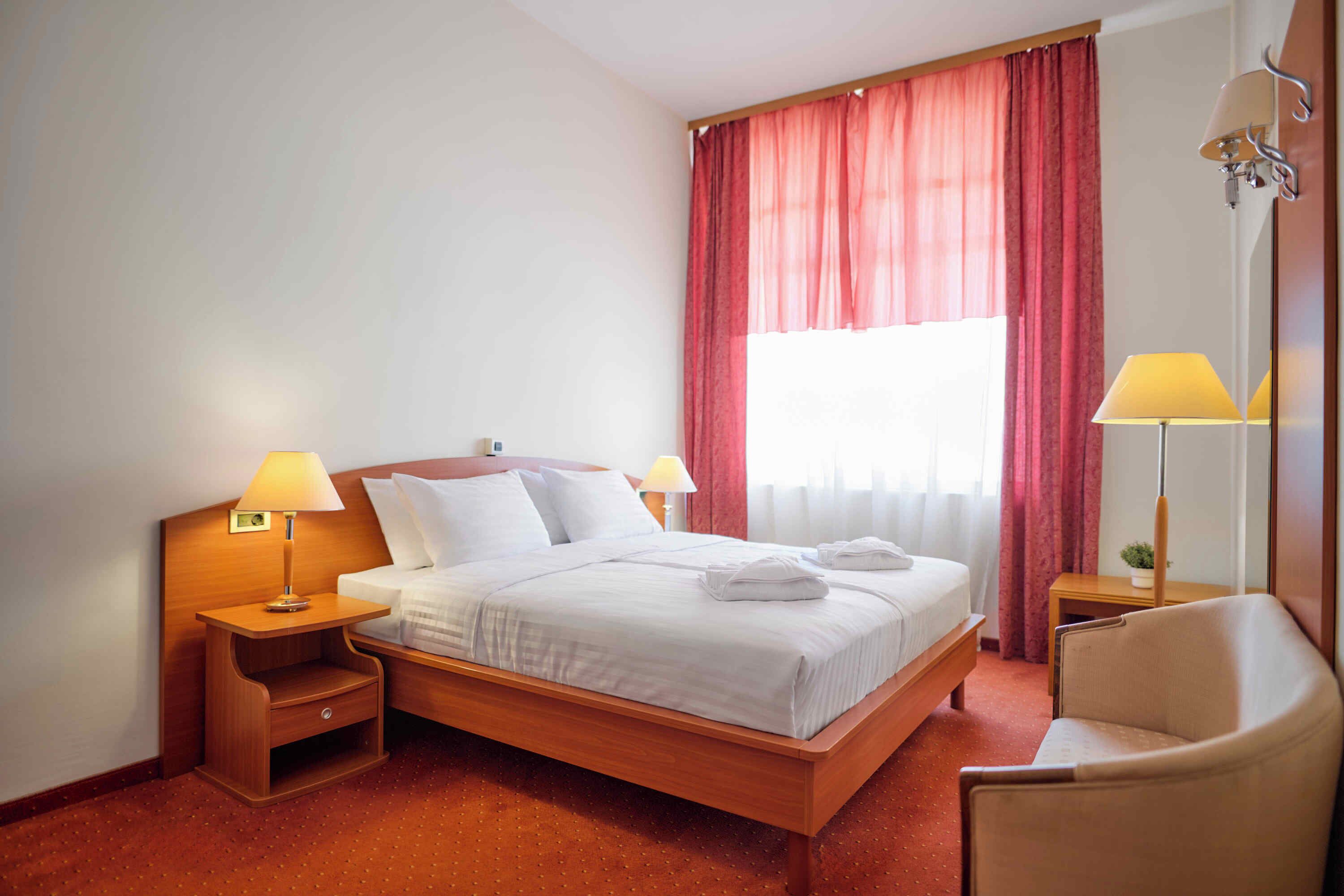 A hotel room with a wooden bed, two bedside tables with lamps, a chair, and a large window with red curtains.