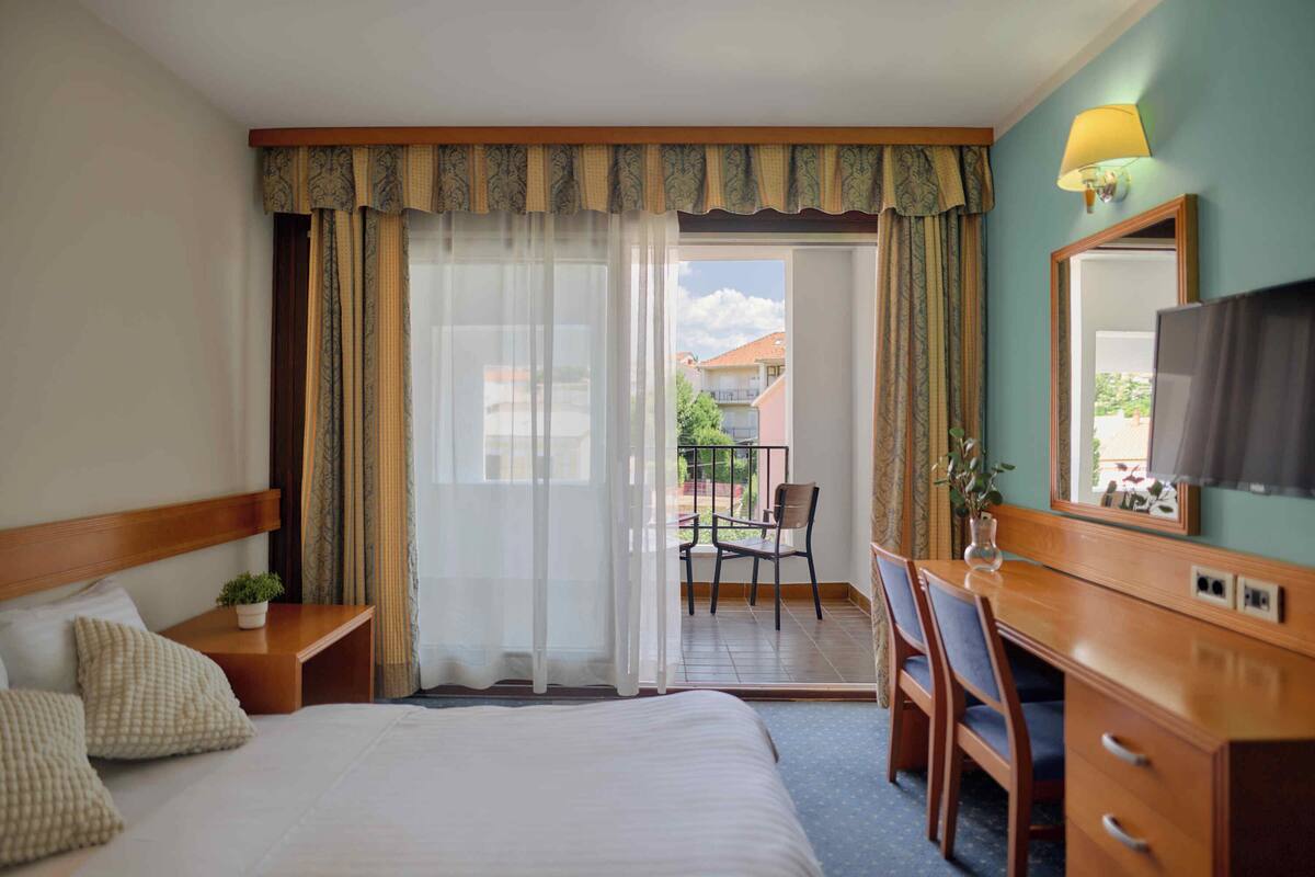 A hotel room with a bed, a desk with a chair, a mirror, and a view of the outside balcony.