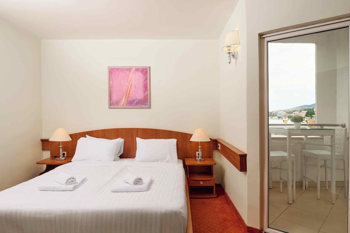 A hotel room with a bed, two bedside tables with lamps, a painting on the wall, and a balcony with chairs.