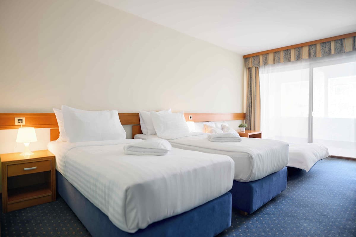 A hotel room with two beds, a nightstand with a lamp, and a large window with curtains.