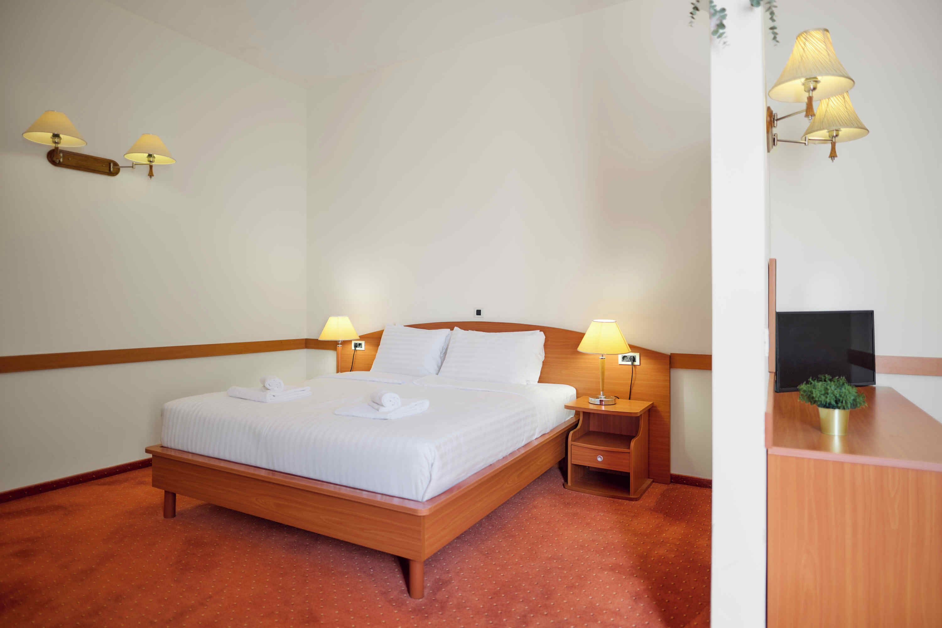 Ein Hotelzimmer mit einem hölzernen Bett, zwei Nachttischlampen, einem Nachttisch aus Holz, einem Schreibtisch mit einer Pflanze und einem Flachbildfernseher.