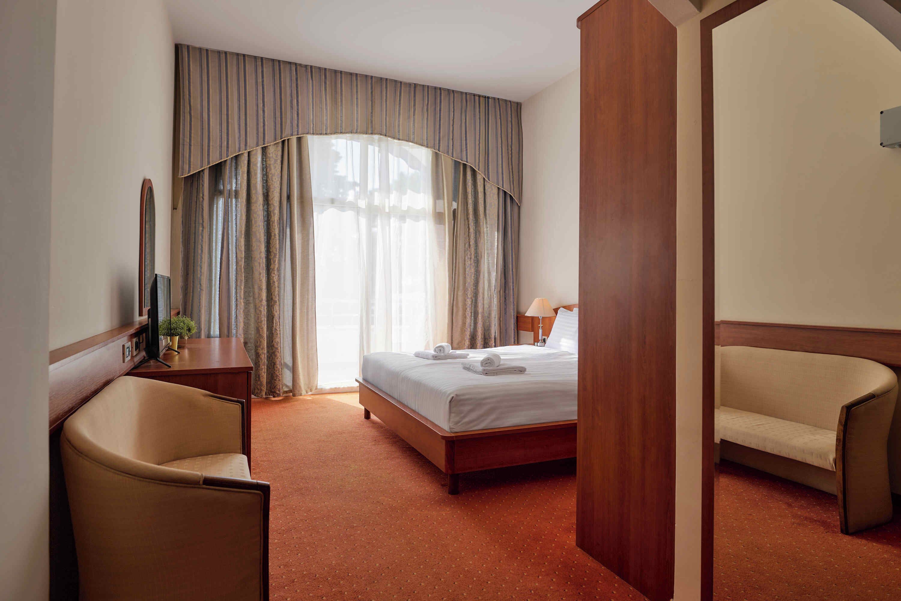 A hotel room with a bed, a chair, a desk, and a large window with curtains.