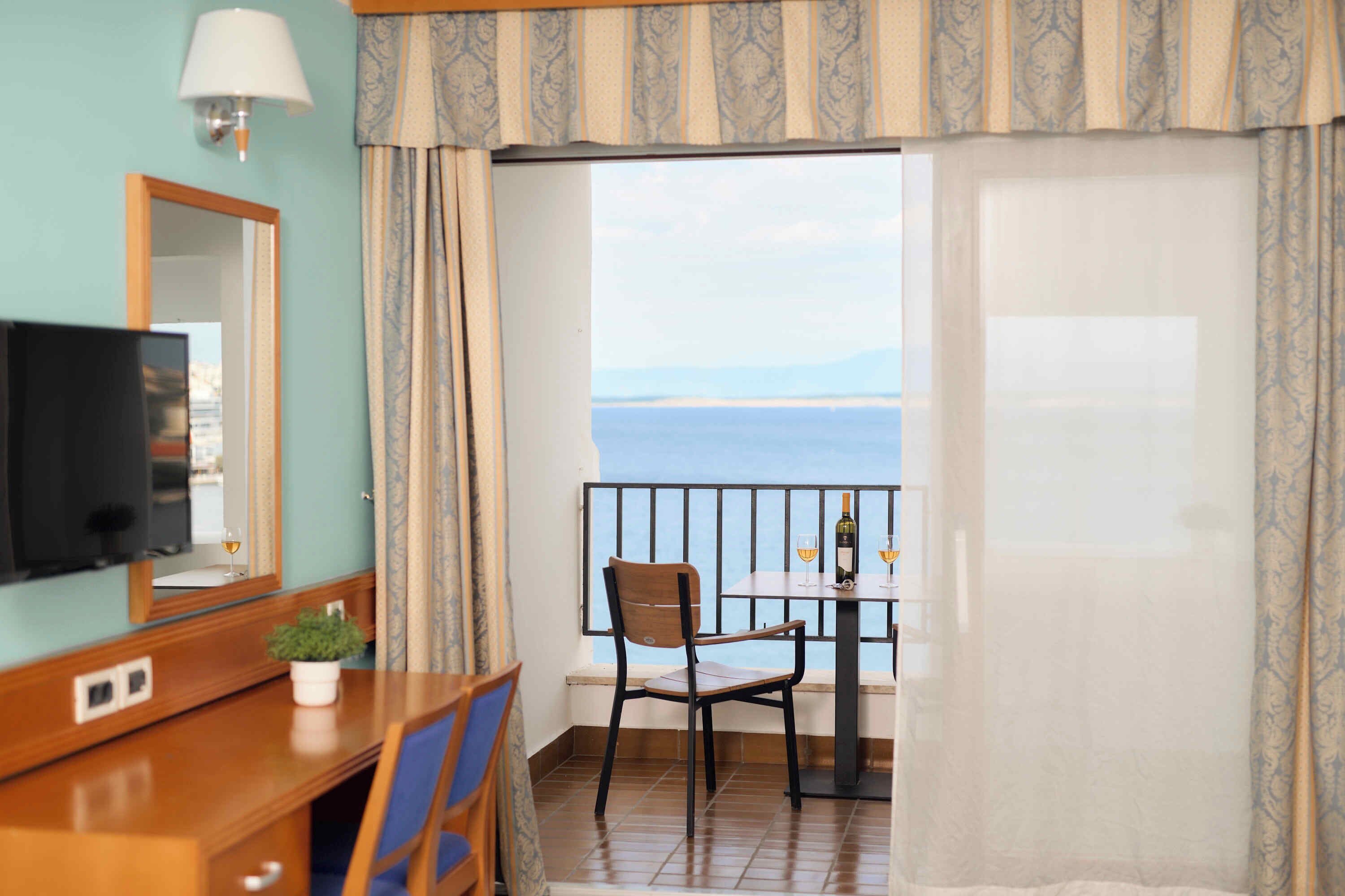 A hotel room with a TV, a small table with a chair, a potted plant, and a view of the sea through a balcony.