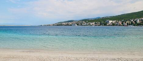 A beach with clear turquoise water, a sandy shore, and a town in the background.