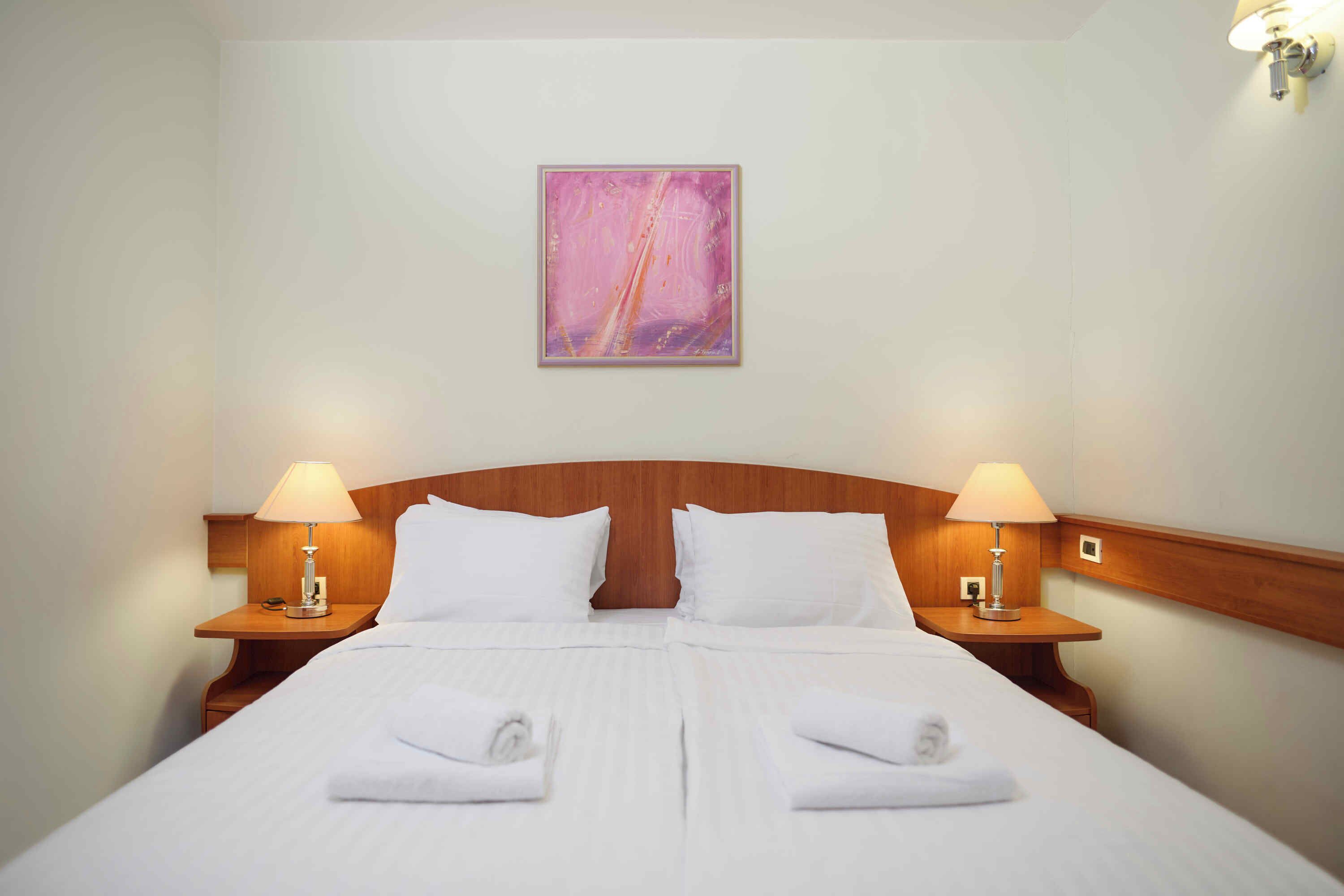 A hotel room with a double bed, two bedside lamps, a wooden headboard, and a painting on the wall.