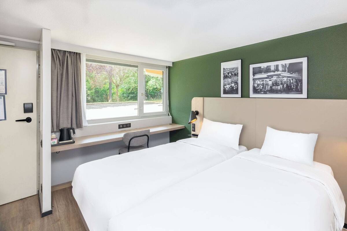 A hotel room with a bed, a desk, a chair, a window with curtains, and two black and white photographs on the wall.