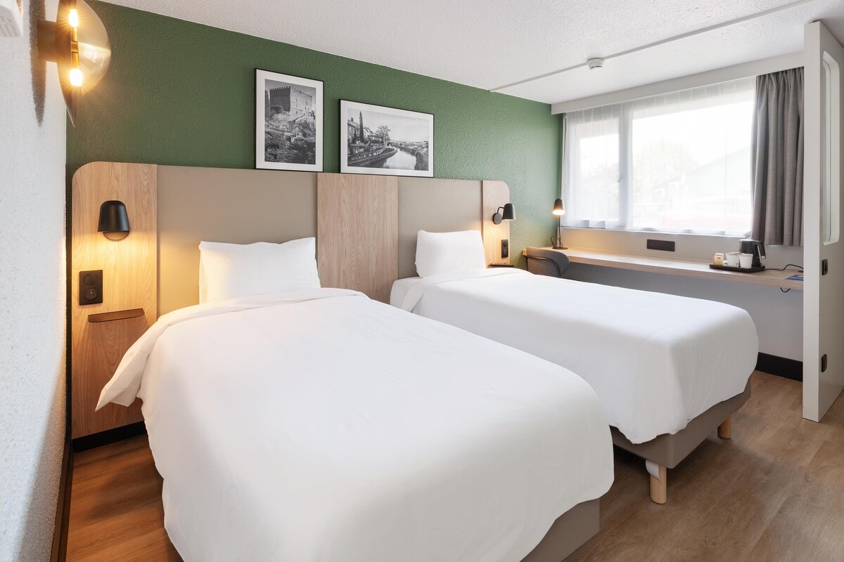 A hotel room with two beds, a desk, a window with curtains, and two framed pictures on the wall.