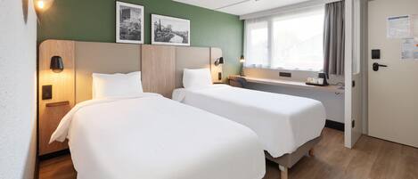 Une chambre d’hôtel avec deux lits, un bureau, une fenêtre avec des rideaux et deux tableaux encadrés au mur.