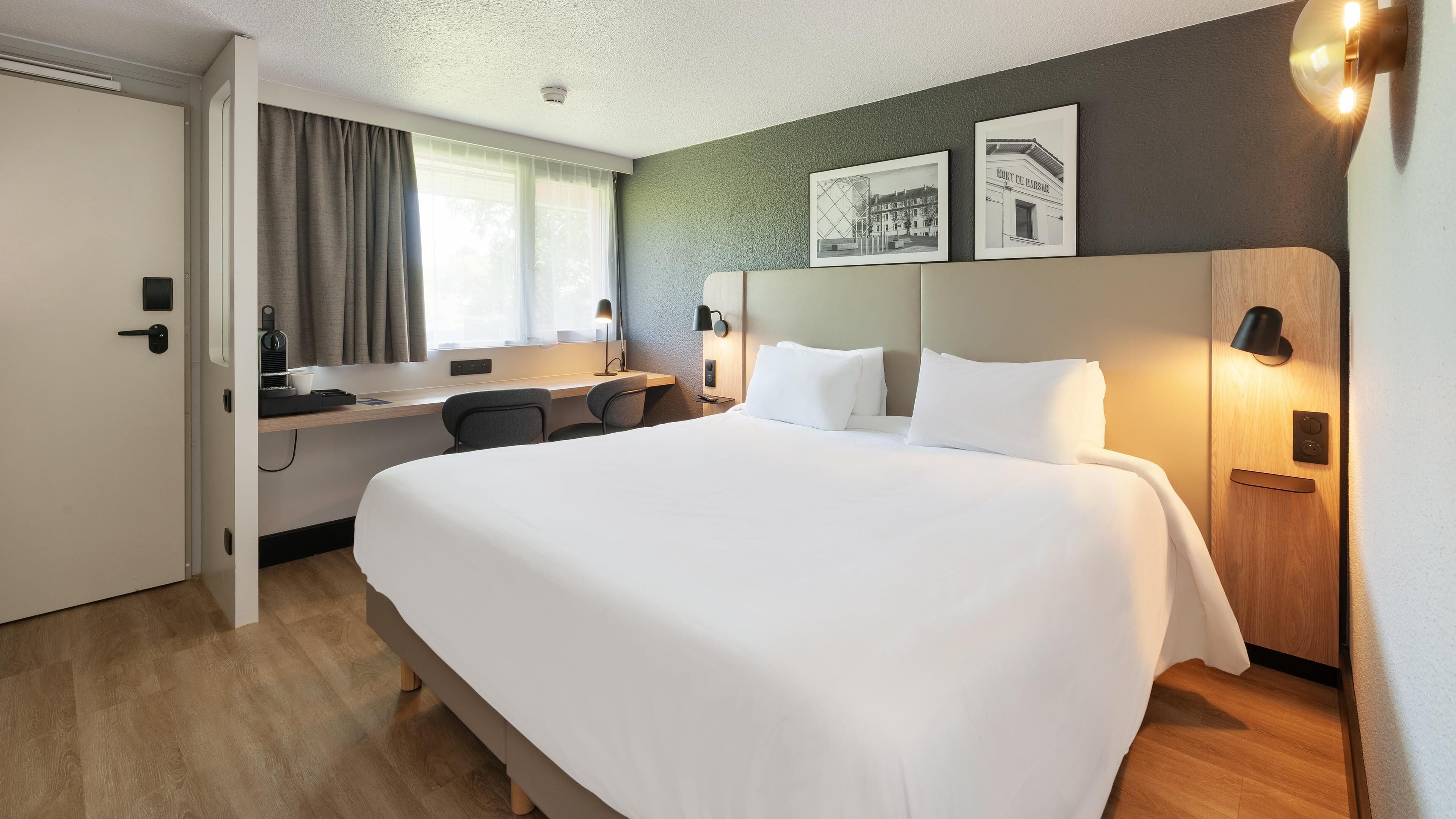 Habitación de hotel con una cama grande, un escritorio con silla, una computadora y un ventanal con cortinas.