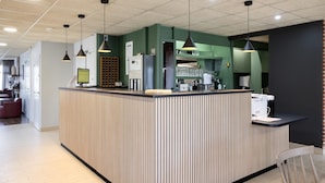 A modern reception area with a wooden reception desk, pendant lighting, and a coffee machine. - Campanile NATURE - Mont de Marsan (Mont-de-Marsan)
