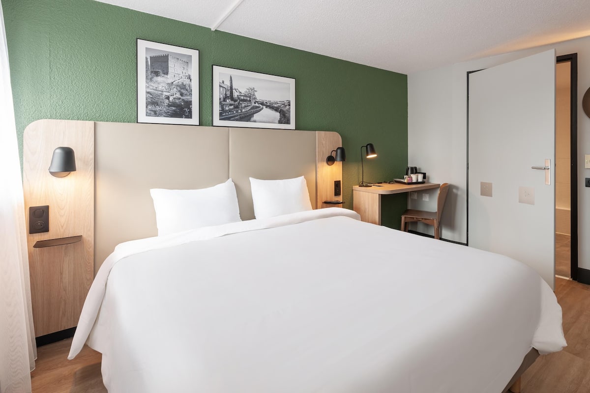A hotel room with a large bed, a desk, a chair, and a wardrobe. There are two framed pictures on the wall above the bed.