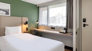 A hotel room with a bed, a desk with a lamp, a coffee maker, and a window with curtains.