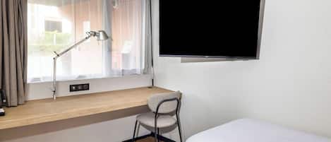 Habitación de hotel con cama, escritorio con silla, televisor montado en la pared y ventana con cortinas.