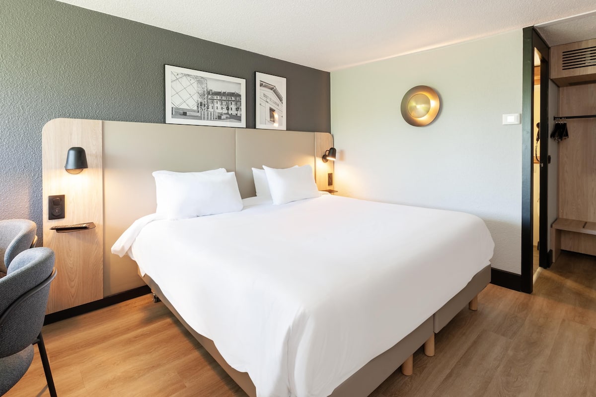 A hotel room with a large bed, a chair, a desk, and a wardrobe.