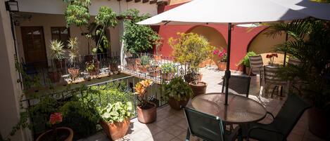 Un patio con una mesa redonda y sillas, rodeado de plantas en macetas y una sombrilla blanca.