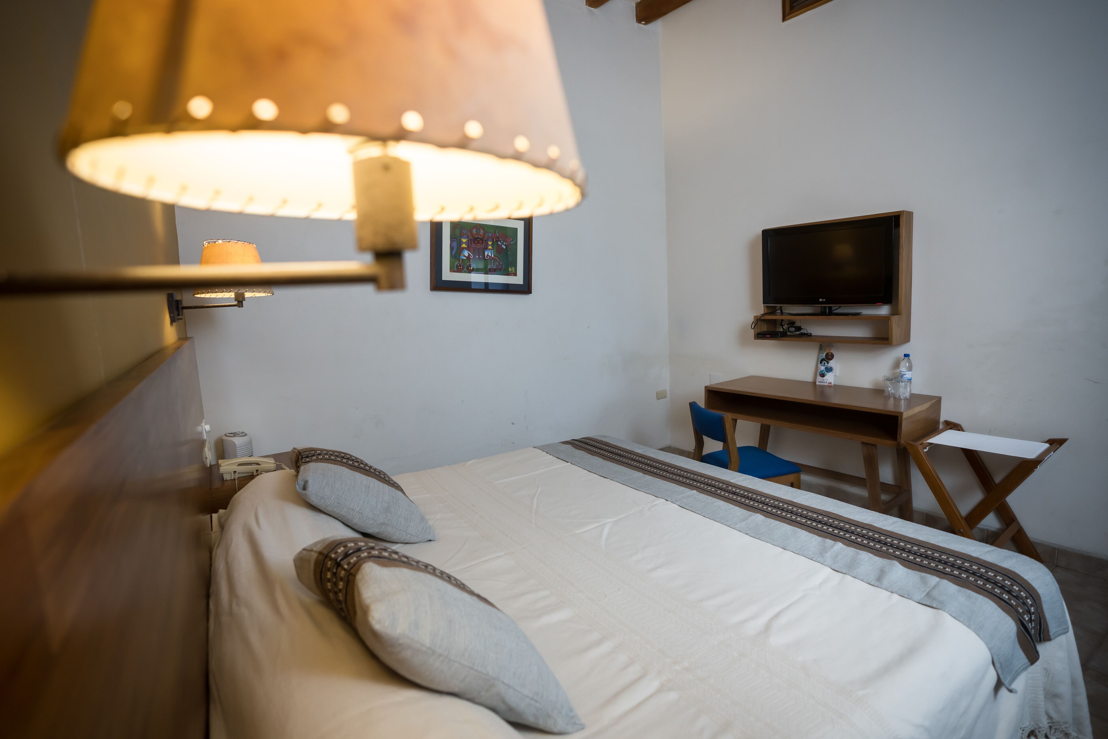 Un lit bien fait, avec des oreillers, une lampe de chevet, un téléviseur posé sur un meuble en bois et un bureau avec une chaise.