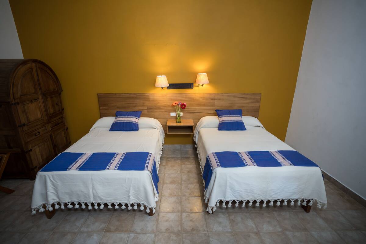 A room with two beds, a wooden wardrobe, a nightstand with lamps, and a vase with flowers.
