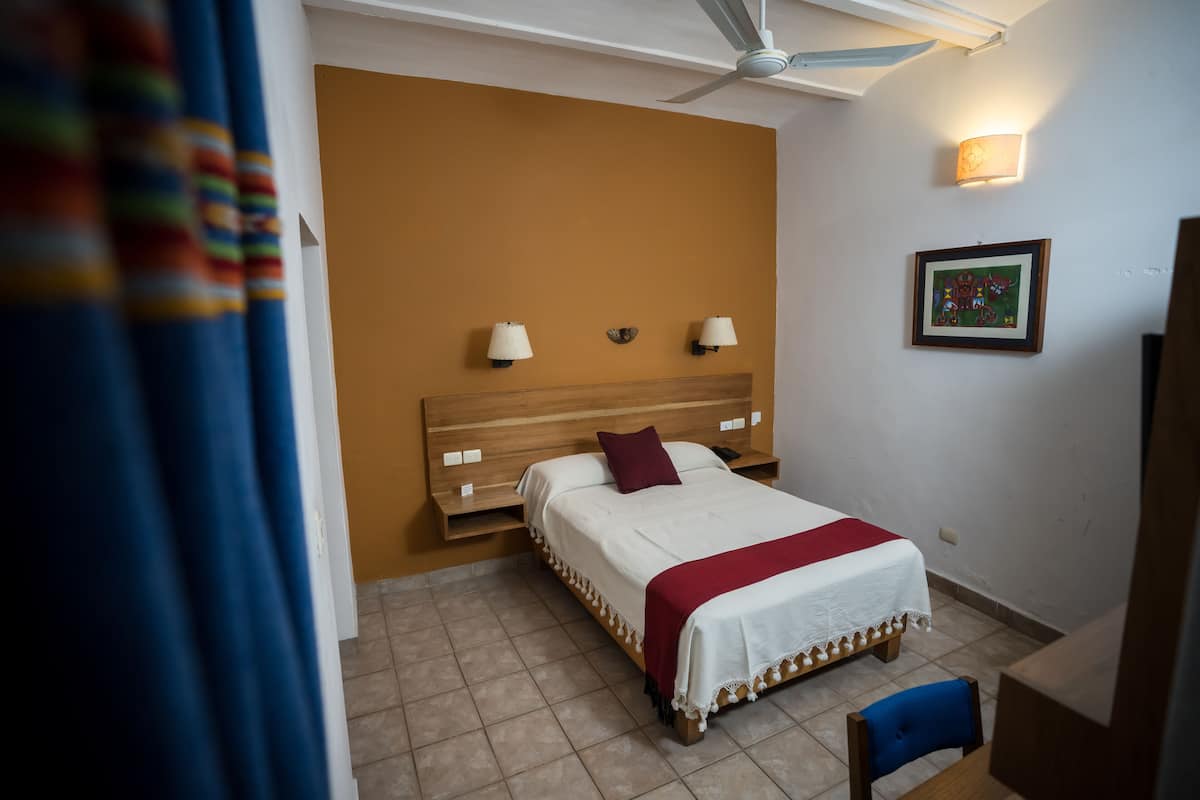 A bedroom with a bed, bedside lamps, a ceiling fan, a framed picture on the wall, and a chair.