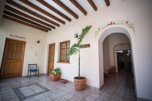 Un pasillo con una puerta de madera que dice "Sta. Margarita", una planta en maceta, una silla y un piso de baldosas.