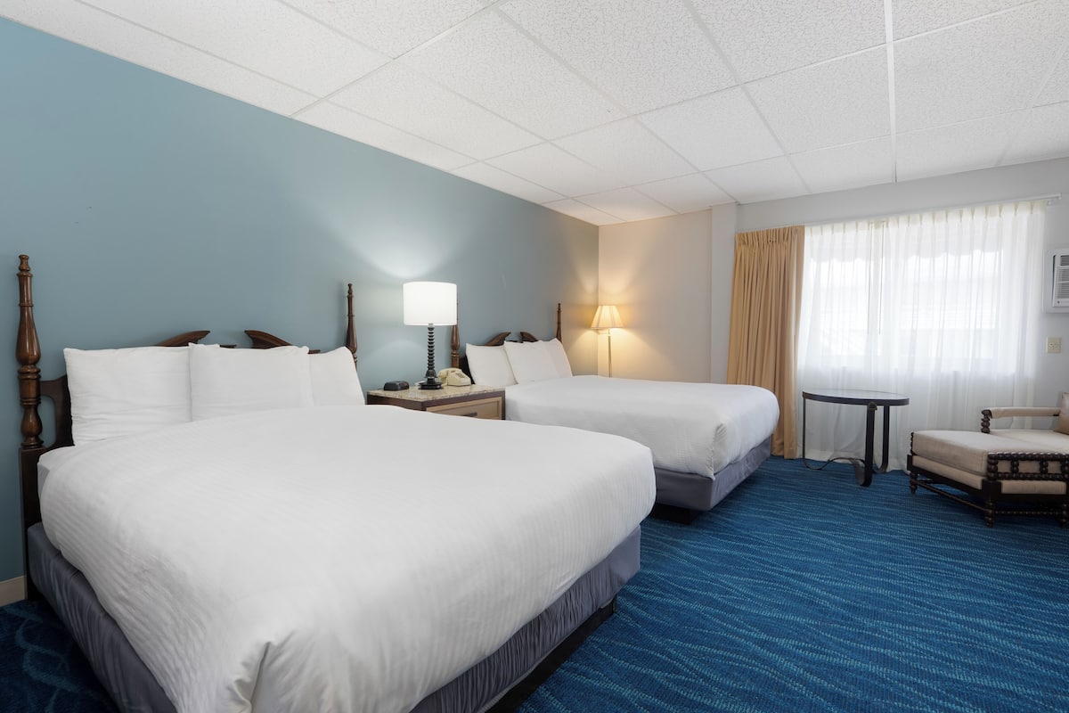 A hotel room with two beds, a small table, a chair, and a window with curtains.
