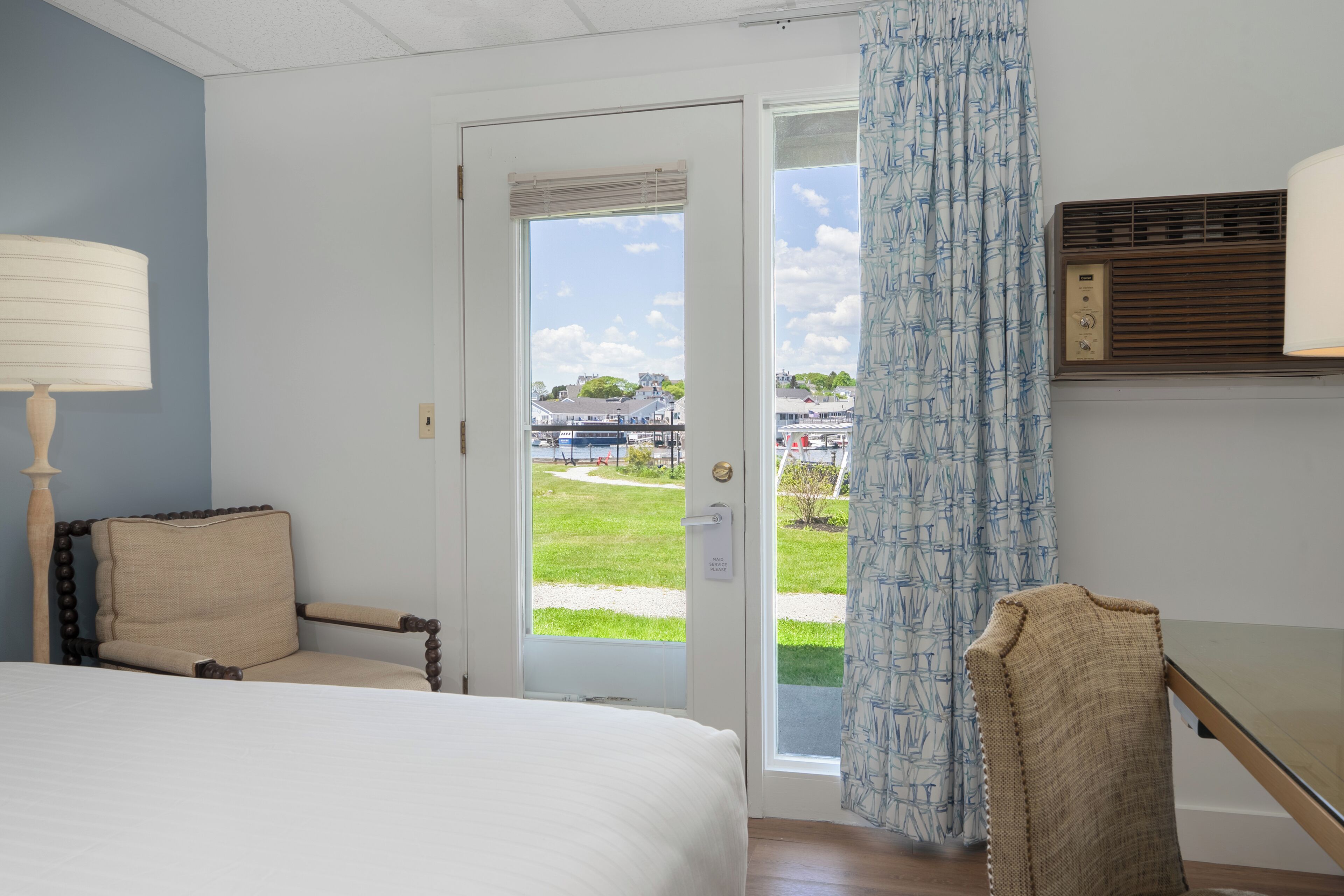 A hotel room with a bed, a chair, a desk, a window with a view of the outdoors, and a wall-mounted air conditioning unit.