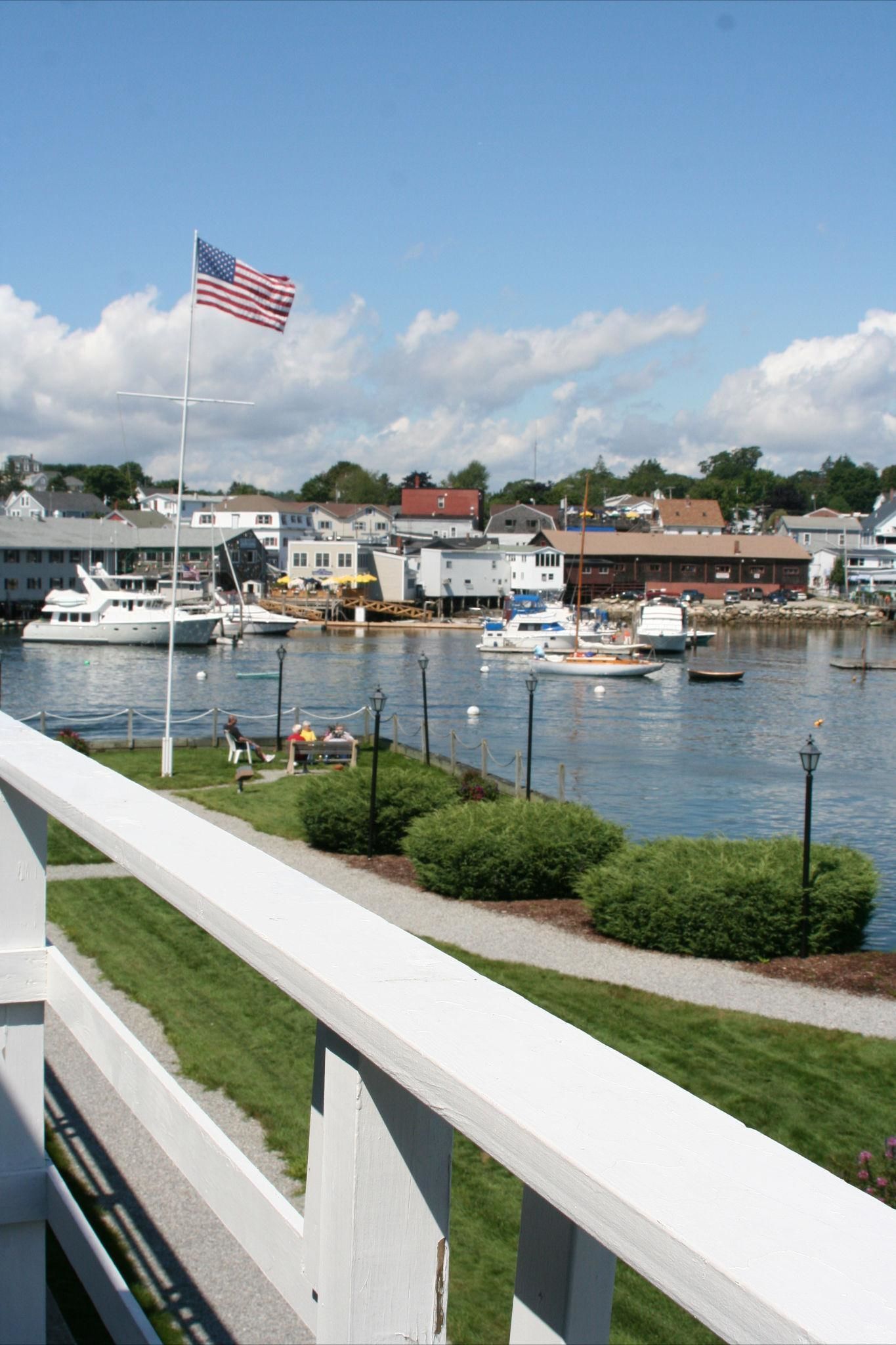A marina with boats docked, a flagpole with an American flag, and buildings in the background.