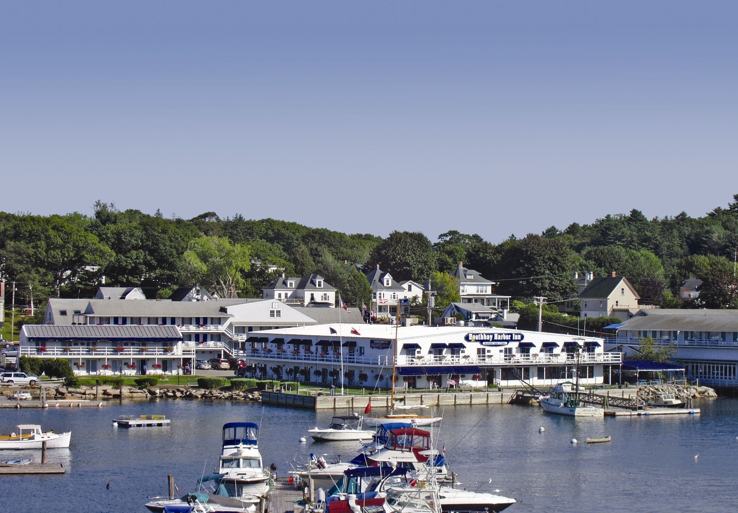 A marina with boats docked, a large building with a blue and white facade, and a forested hill in the background.