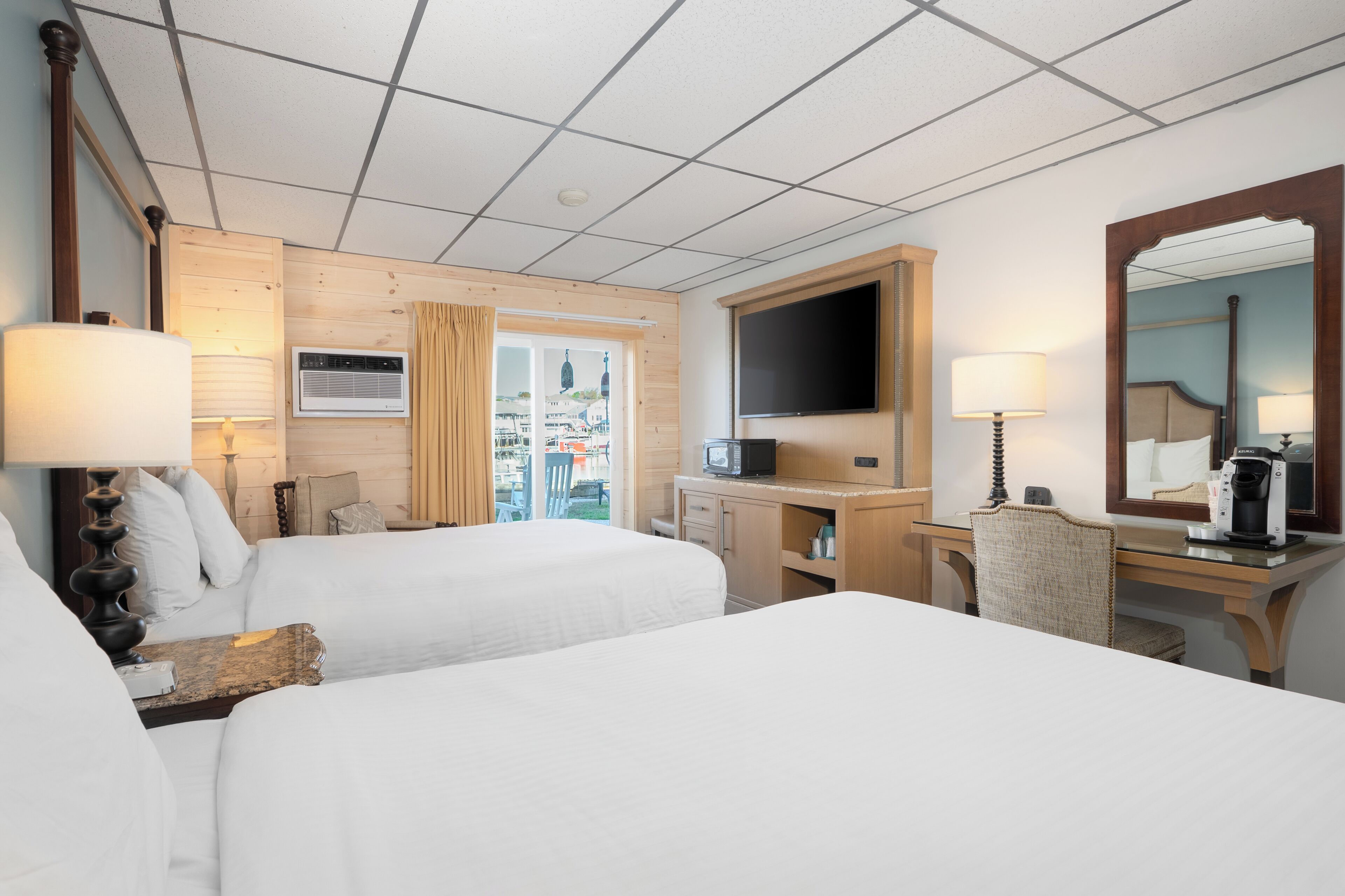 A hotel room with two beds, a desk, a TV, and a window with a view of buildings.