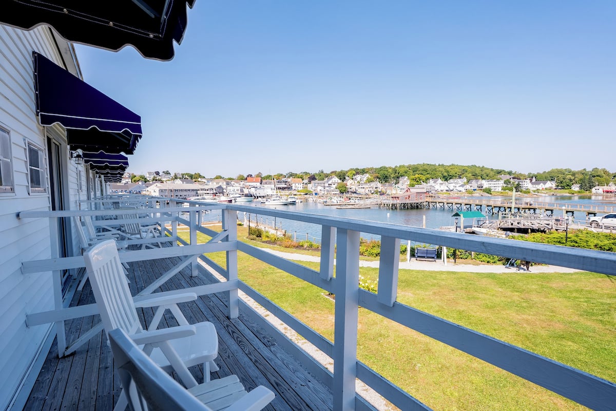 A wooden deck with white chairs and a railing overlooking a marina with boats and a town in the background.