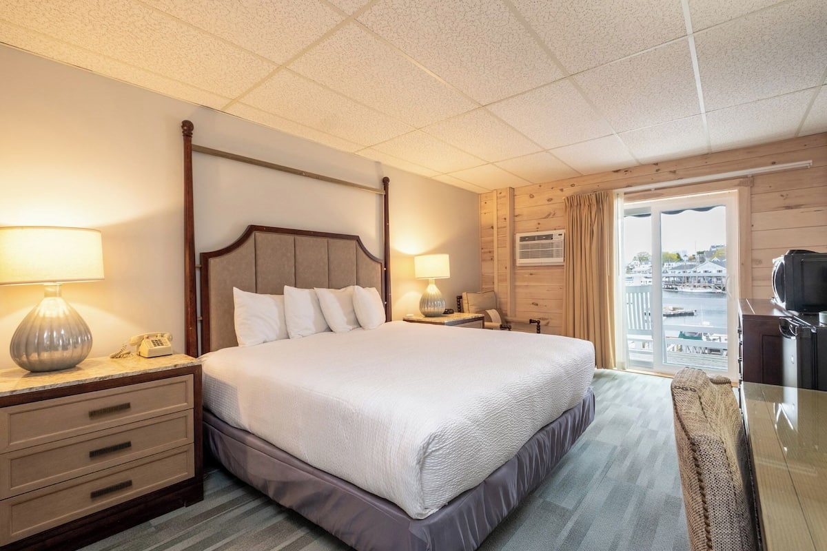 A hotel room with a bed, bedside tables, a lamp, a telephone, a window with a view of the water, and a wooden wall.