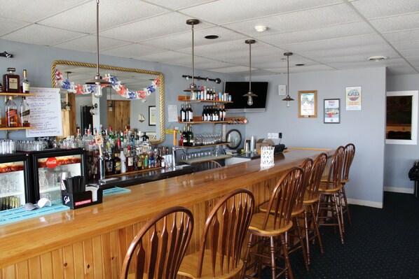 A bar area with wooden bar counter, multiple chairs, shelves with bottles, and a mirror.