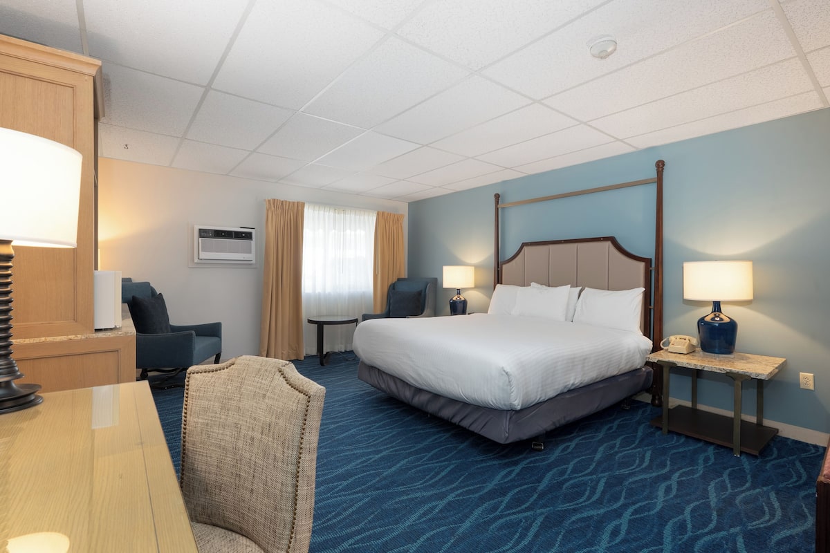 A hotel room with a large bed, two bedside tables with lamps, a desk, a chair, a small table, and a window with curtains.