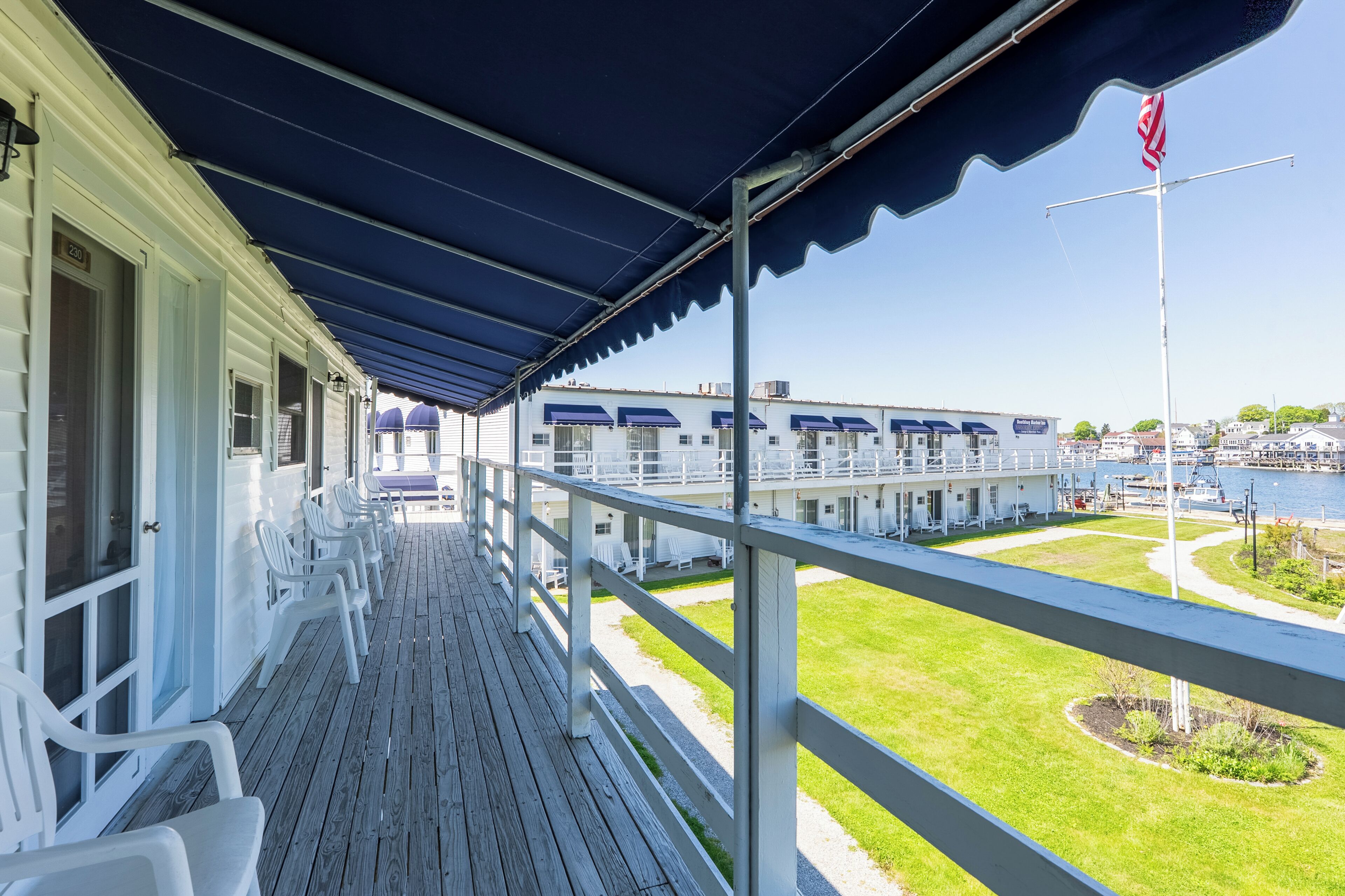 A covered balcony with white chairs, a view of a building with multiple windows, and a flagpole with an American flag.
