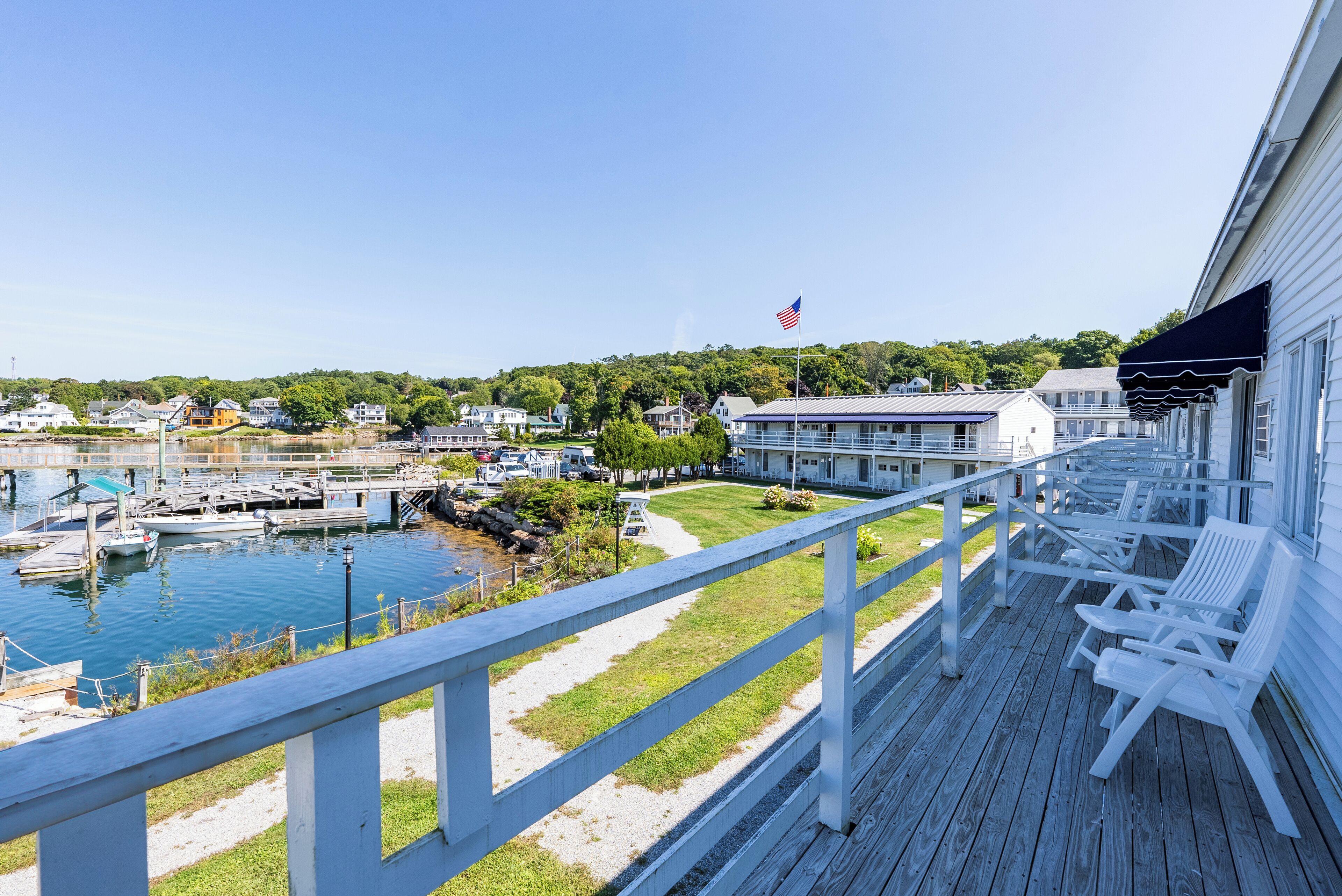 A wooden deck with white chairs overlooking a marina with boats, a grassy area, and a building with an American flag.
