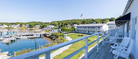 A wooden deck with white chairs overlooking a marina with boats, a grassy area, and a building with an American flag.