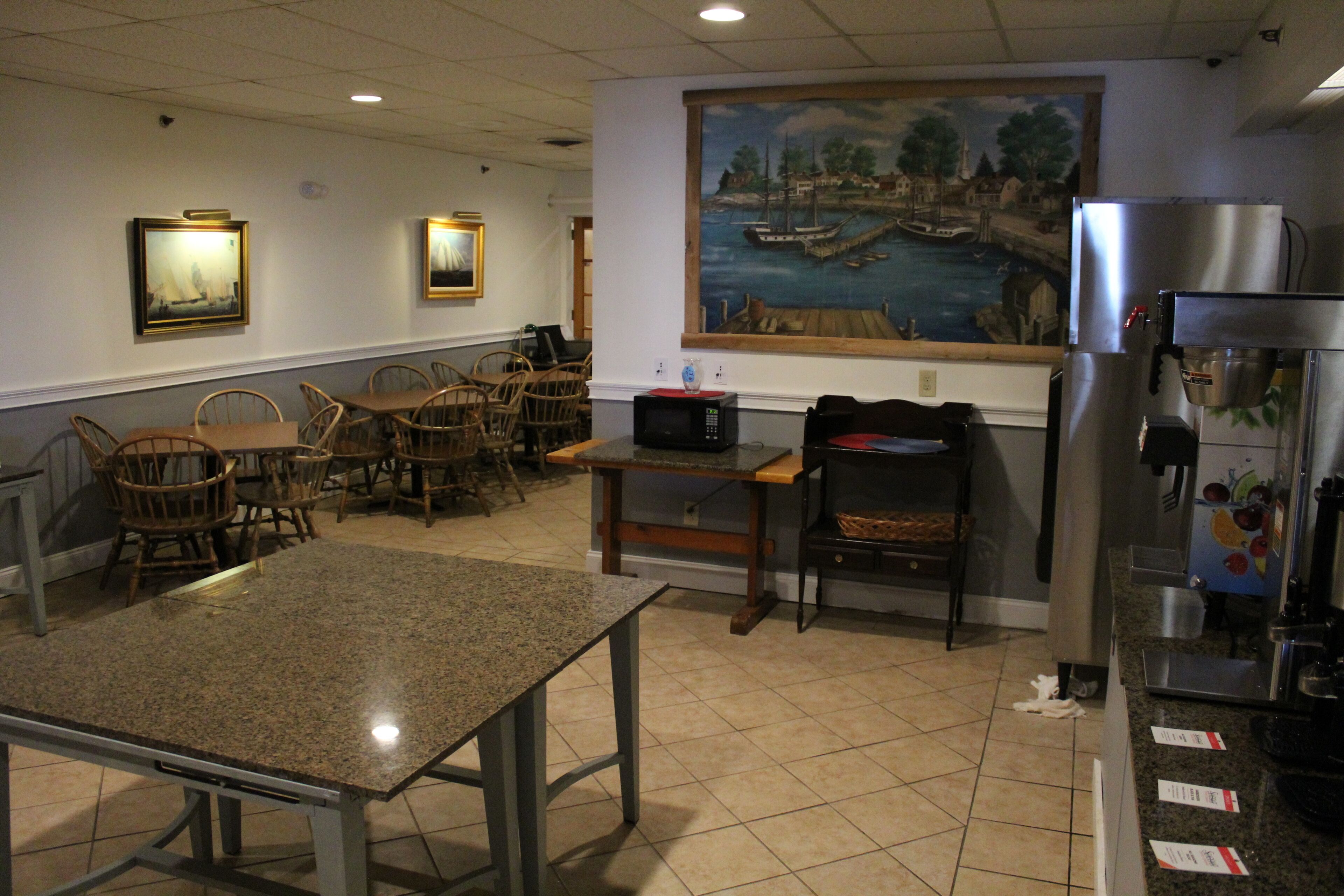 A restaurant with a dining area featuring tables and chairs, a kitchen area with a refrigerator and coffee machine, and a mural on the wall depicting a harbor scene.