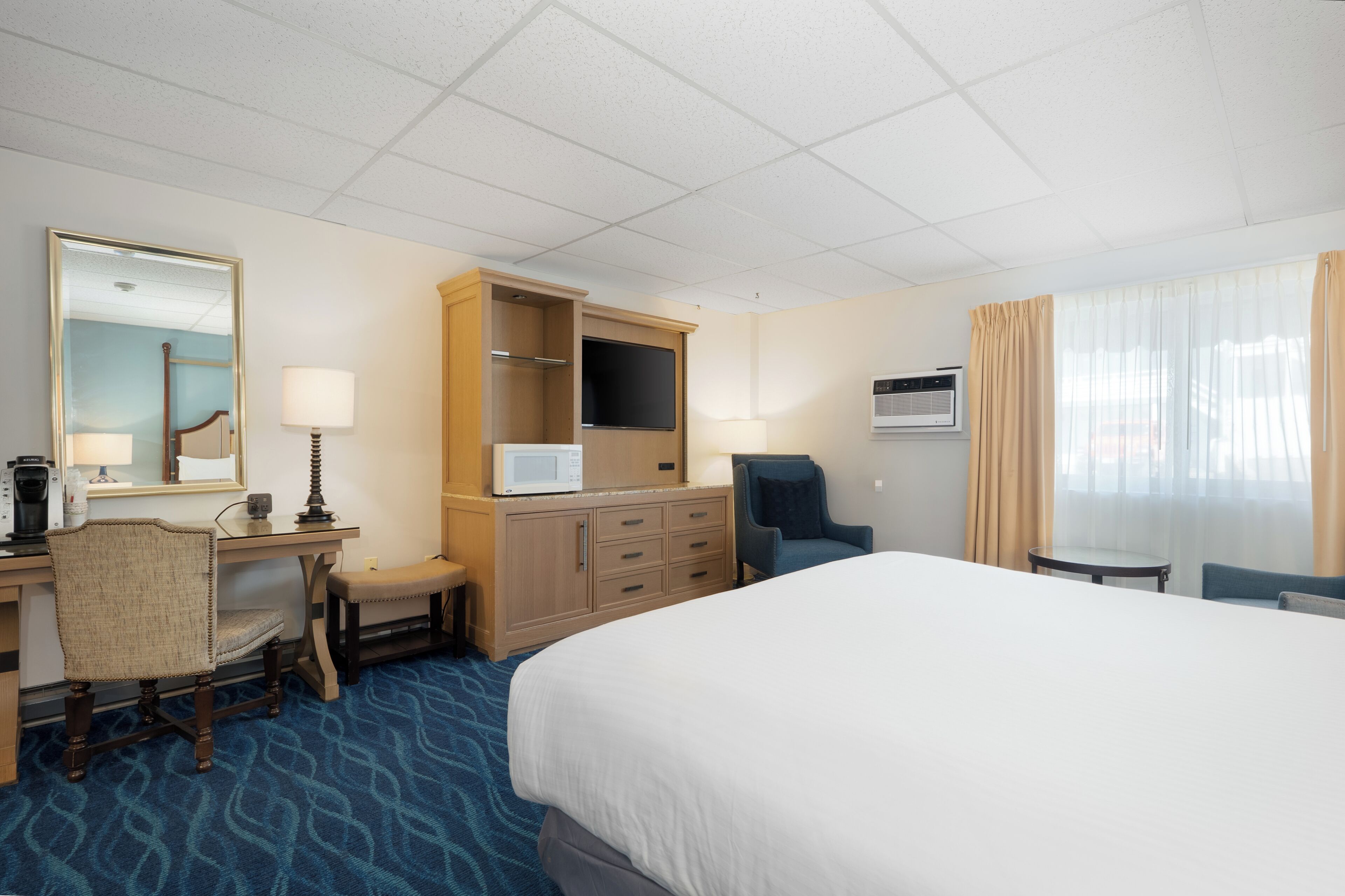 A hotel room with a large bed, a desk with a chair, a television, a minibar, and a window with curtains.