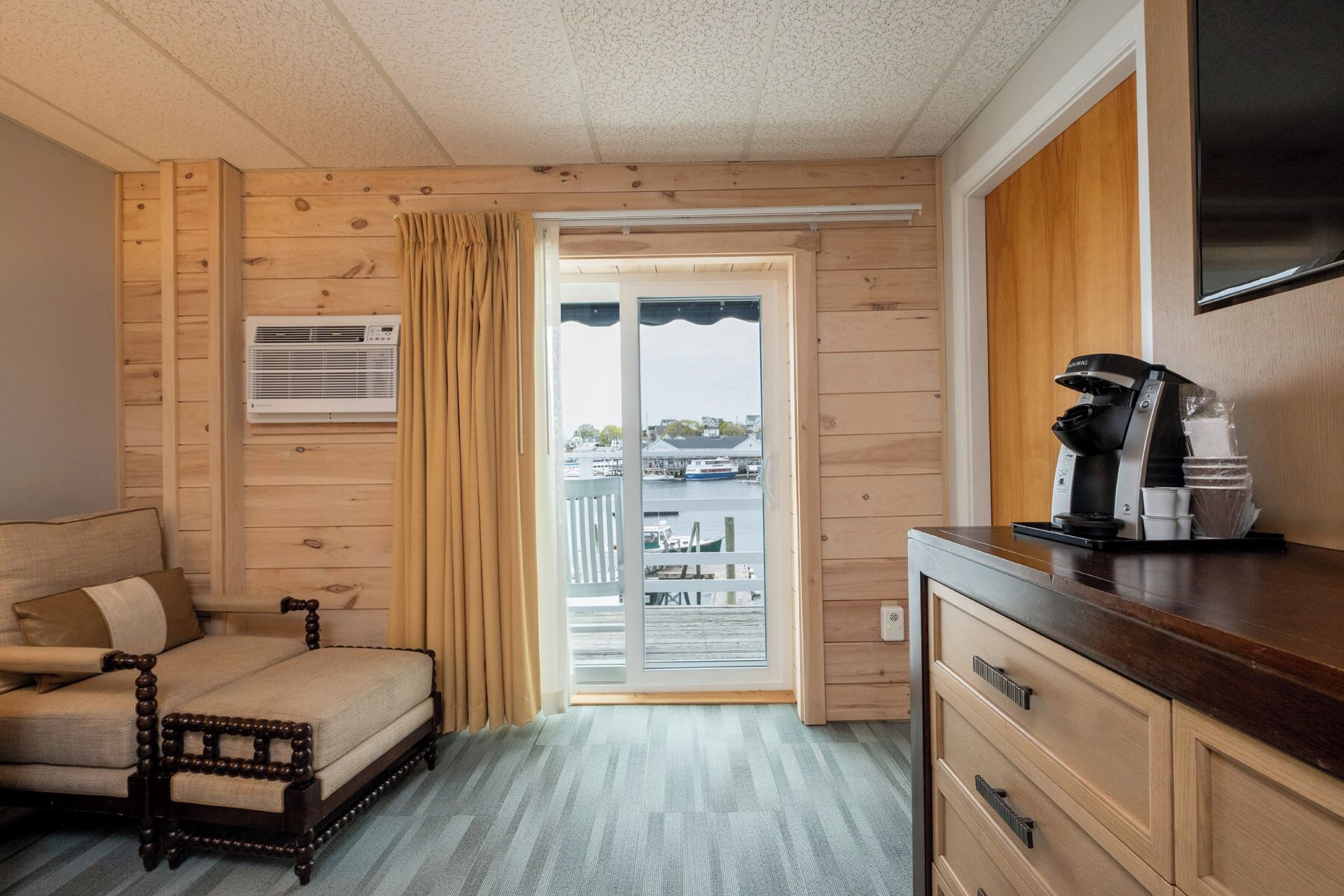 Une pièce avec un intérieur en bois, un canapé, une machine à café et une vue sur une marina, visible à travers une porte vitrée.