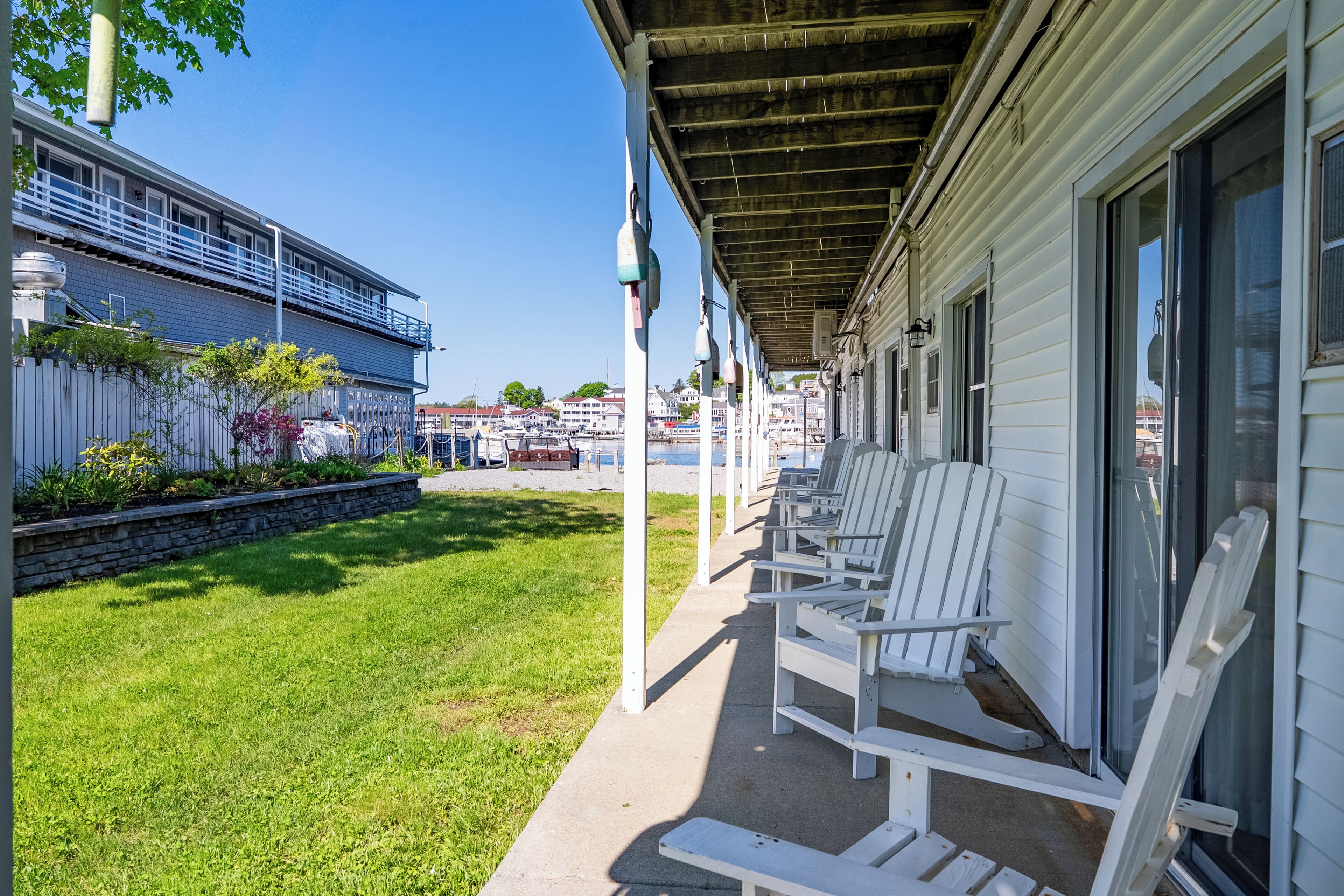 Een overdekt terras met witte Adirondack-stoelen, een stenen steunmuur en een uitzicht op een jachthaven met boten en gebouwen op de achtergrond.