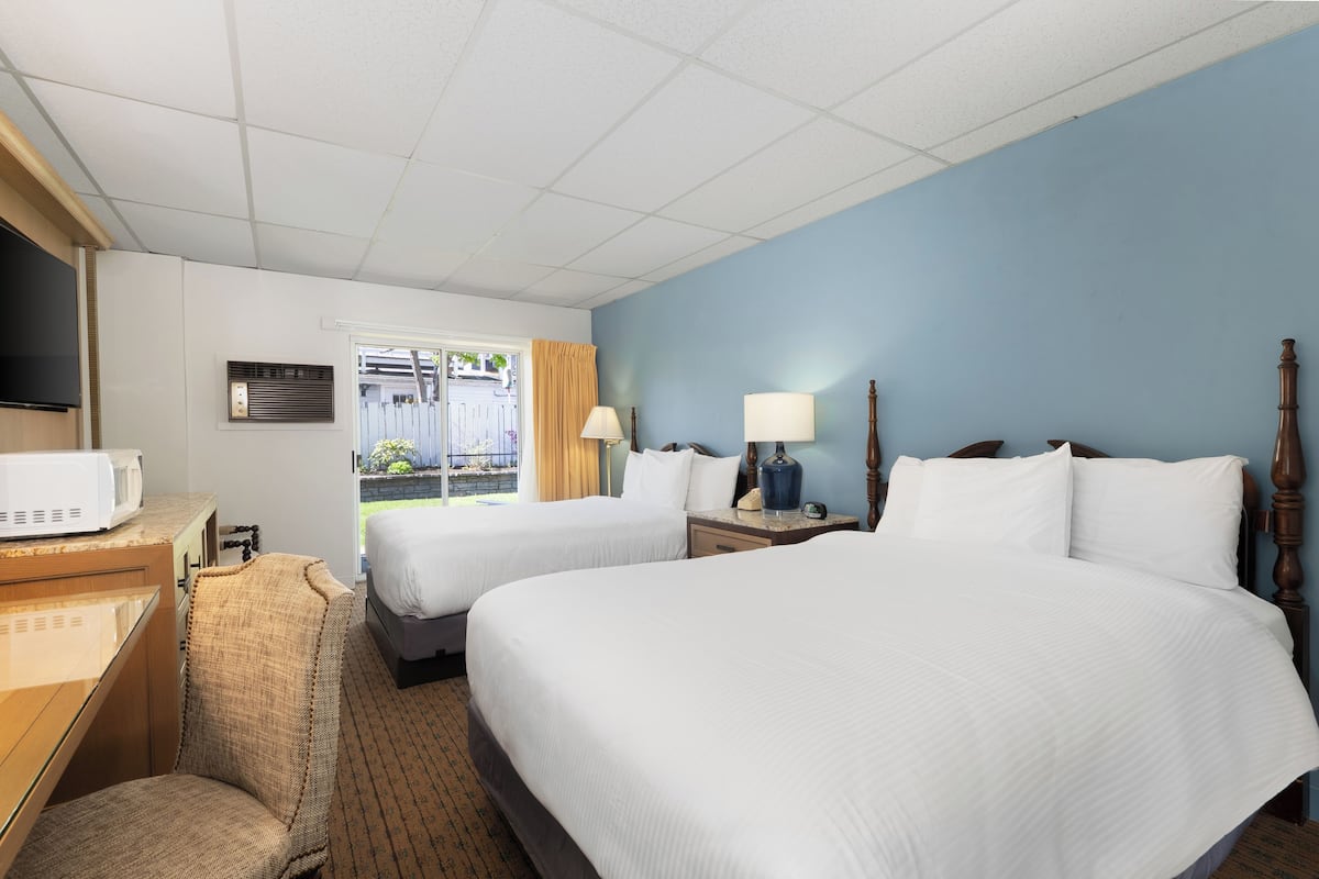 A hotel room with two beds, a desk with a microwave, a chair, a TV, a window with curtains, and a lamp.