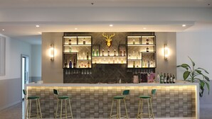 Zona de bar con mostrador de mármol, varias estanterías llenas de botellas de licor y tres taburetes verdes.